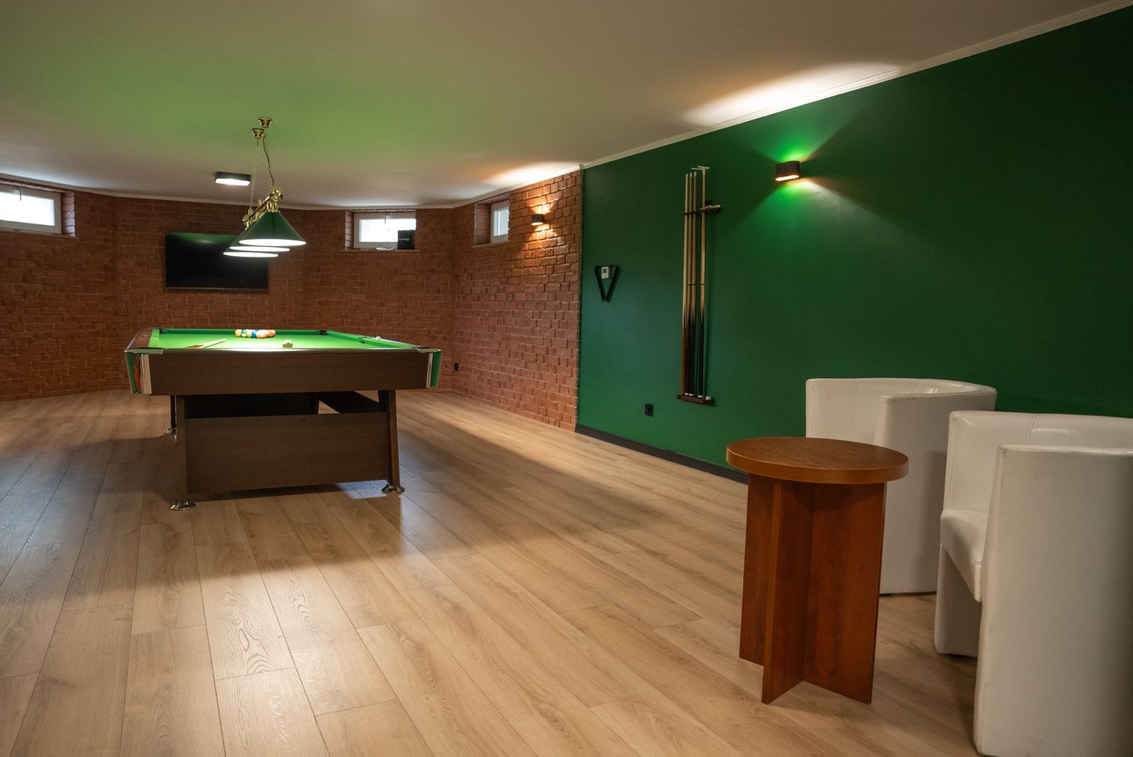 Pokój z ceglanymi ścianami i zieloną ścianą, wyposażony w stół bilardowy, drewniany stolik i białe krzesła, oświetlony lampami z zielonymi abażurami.