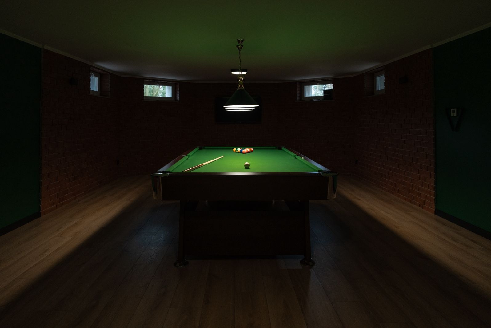 Stół bilardowy z zielonym suknem, ustawionymi bilami i kijem, oświetlony wiszącą lampą, umieszczony w pomieszczeniu z ceglanymi ścianami i małymi oknami.