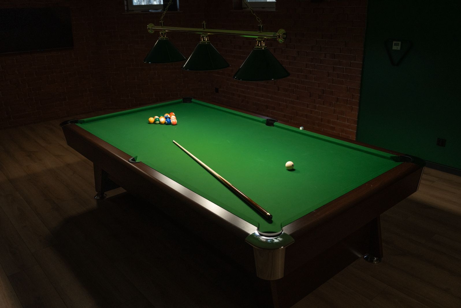 Stół bilardowy z zielonym suknem, ustawionymi bilami i kijem, oświetlony trzema lampami w pomieszczeniu z ceglaną ścianą.