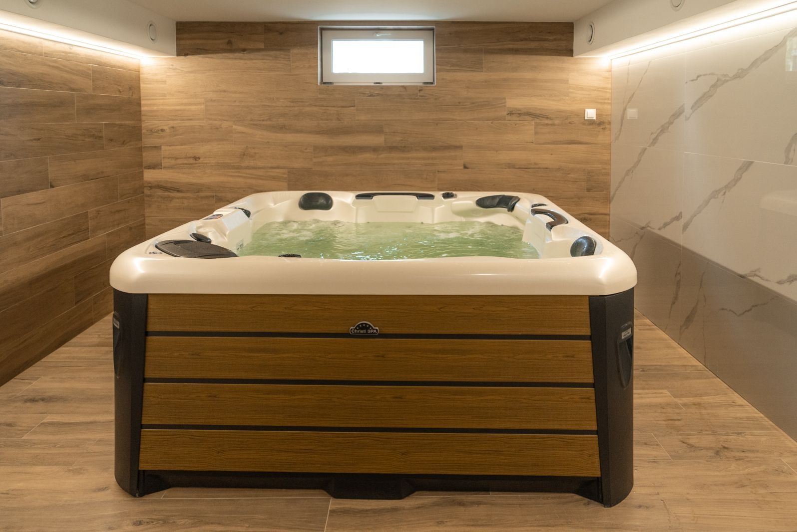 Nowoczesna wanna SPA z hydromasażem, obudowana drewnopodobnymi panelami, wypełniona wodą, w pomieszczeniu wyłożonym płytkami imitującymi drewno i marmur.