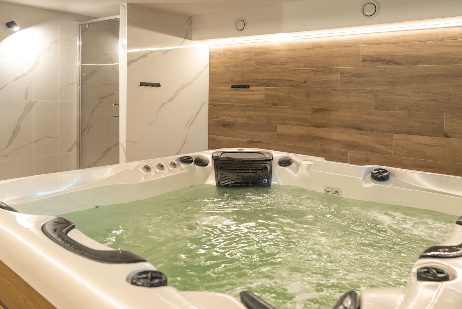 Wnętrze łazienki z dużą, białą wanną jacuzzi wypełnioną wodą, widoczny prysznic ze szklanymi drzwiami i ściany wyłożone białymi i drewnopodobnymi płytkami.