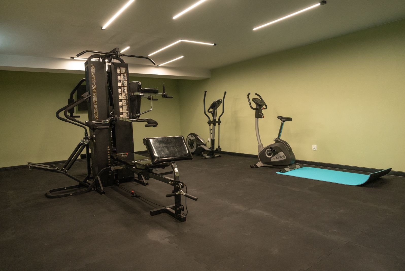 Wnętrze siłowni z wielofunkcyjnym atlasem, orbitrekami i rowerkiem stacjonarnym na czarnej gumowej podłodze, ściany w kolorze stonowanej zieleni, oświetlenie sufitowe w postaci podłużnych lamp LED.