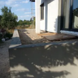 Wczesny etap budowy domu: widoczny fundament z betonu, piasek, ściana budynku z oknem oraz rusztowanie w tle.