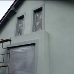 Fragment elewacji budynku z oknami i drzwiami zabezpieczonymi folią malarską, widoczne rusztowanie po lewej stronie, świeżo nałożony tynk w odcieniach szarości.