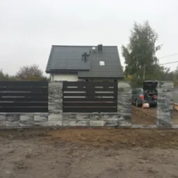 Nowoczesne ogrodzenie z poziomymi, grafitowymi przęsłami i kamiennymi słupkami w odcieniach szarości, na tle domu z panelami słonecznymi na dachu, w trakcie montażu, z widocznym samochodem...