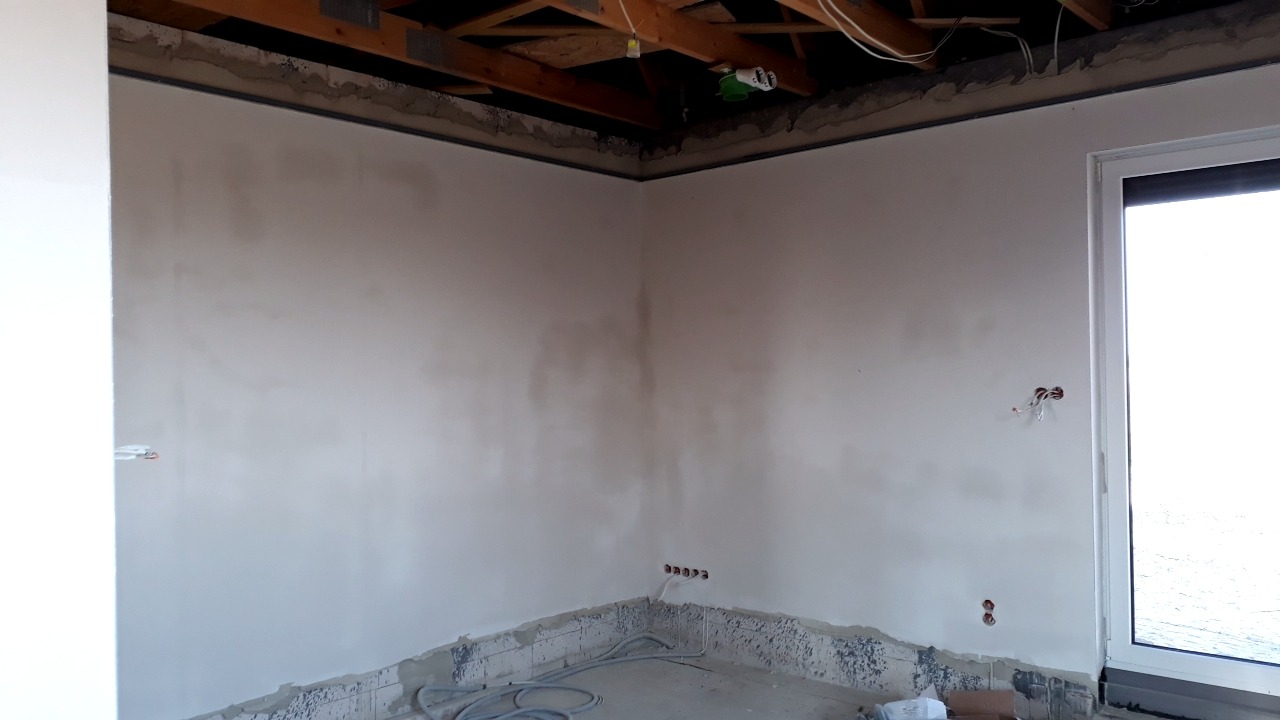 Wnętrze w trakcie prac wykończeniowych: surowa ściana z widocznymi przewodami elektrycznymi, fragment drewnianej konstrukcji stropu i okno z widokiem na zewnątrz.