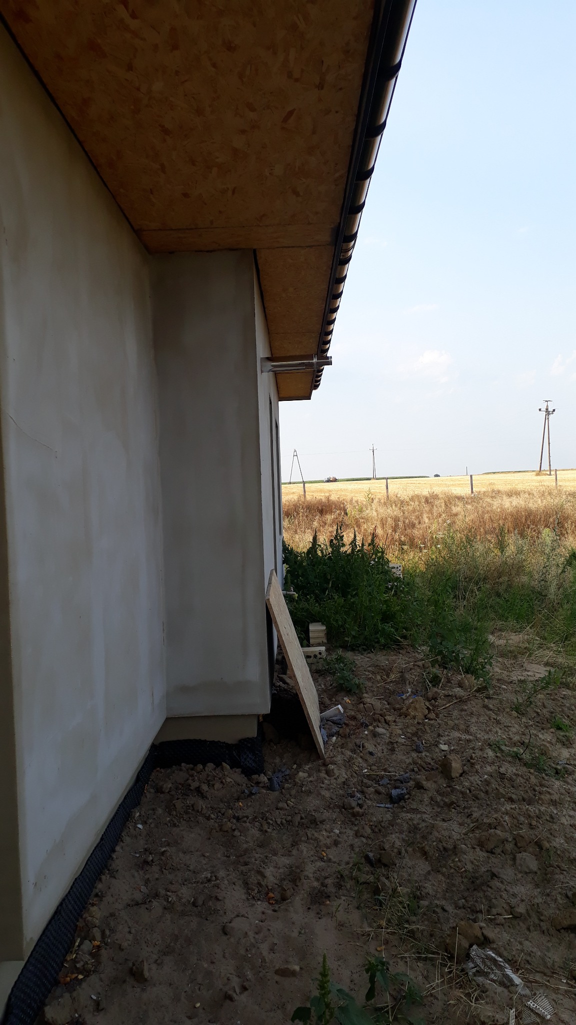Narożnik budynku w trakcie budowy, widoczna surowa elewacja, płyta OSB pod dachem, rynna, folia fundamentowa i niezagospodarowany teren z chwastami w tle.