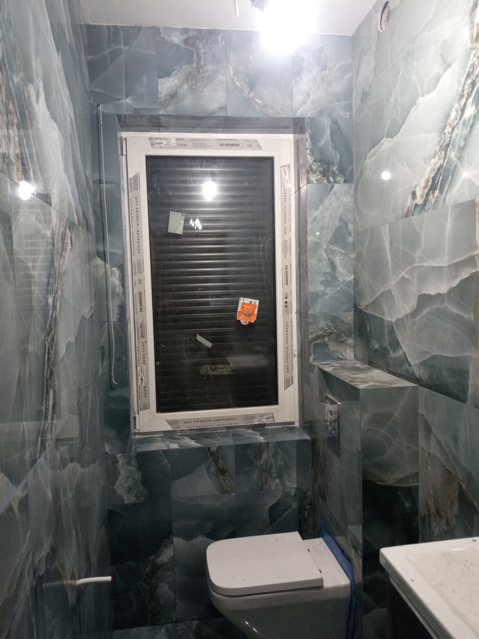 Wykończona łazienka z płytami imitującymi marmur w odcieniach szarości i błękitu, widoczne okno z żaluzjami, toaleta i fragment umywalki, pomieszczenie w trakcie sprzątania po remoncie, na oknie...