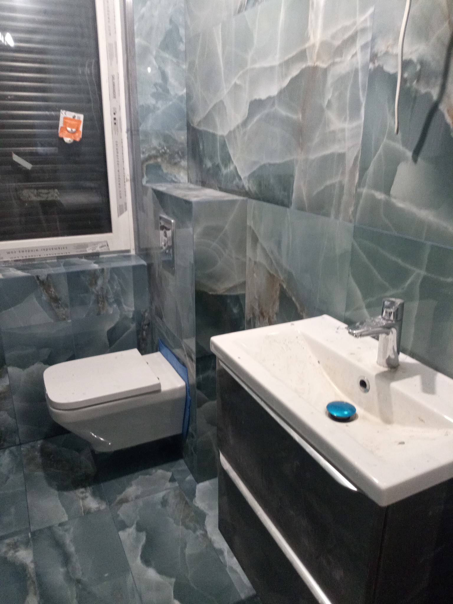 Wykończona łazienka z marmurowymi płytkami w odcieniach zieleni i szarości, wisząca toaleta i umywalka z szafką w ciemnym drewnie, widoczny fragment okna z żaluzjami.