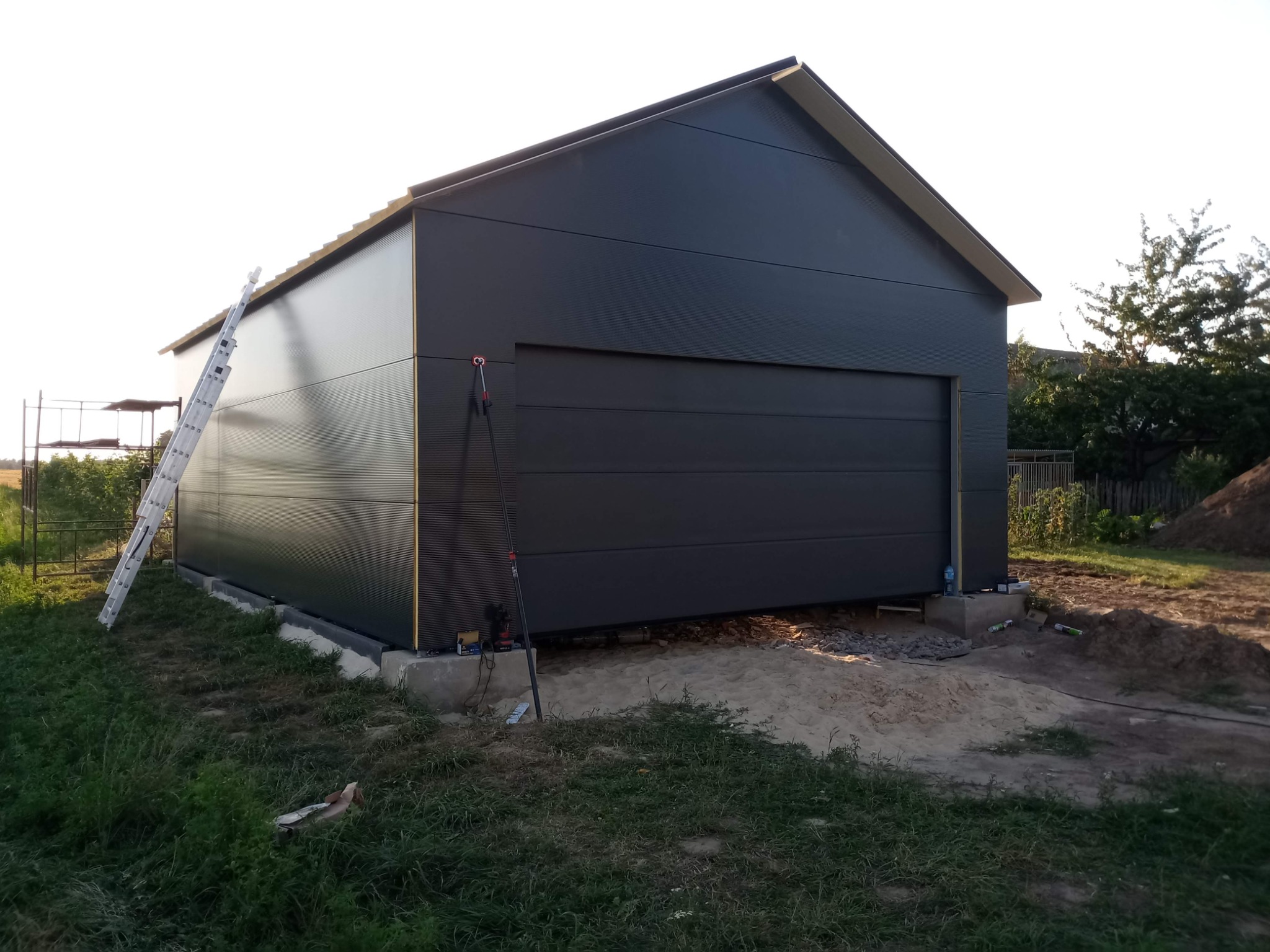 Ciemnoszary budynek z paneli z bramą garażową w trakcie montażu, widoczna drabina oparta o ścianę i narzędzia budowlane u podstawy, otoczenie w stanie surowym.