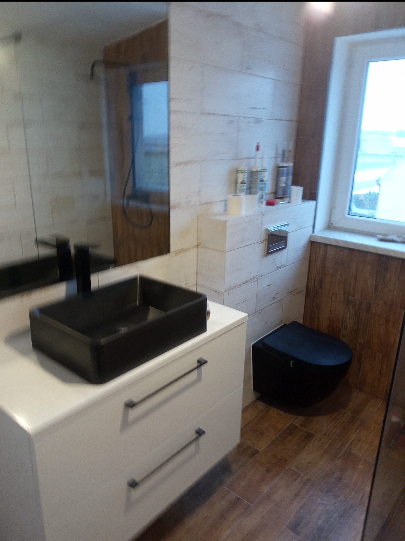 Nowoczesna łazienka z czarną, kwadratową umywalką nablatową, wiszącą toaletą i płytkami imitującymi drewno na ścianach i podłodze.