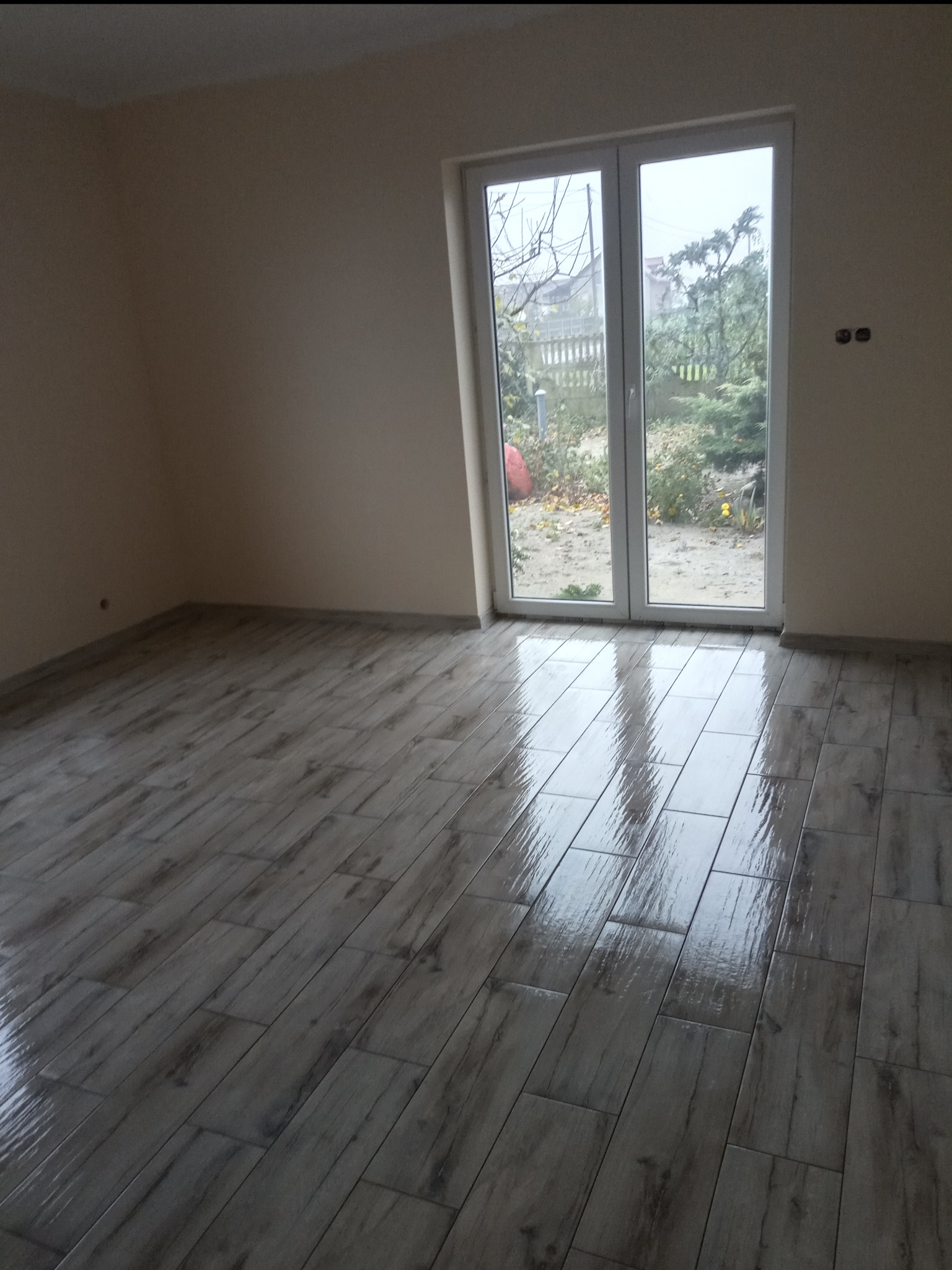 Pusty pokój z nową, błyszczącą podłogą z płytek imitujących drewno, jasne ściany i okno balkonowe z widokiem na ogród w pochmurny dzień.