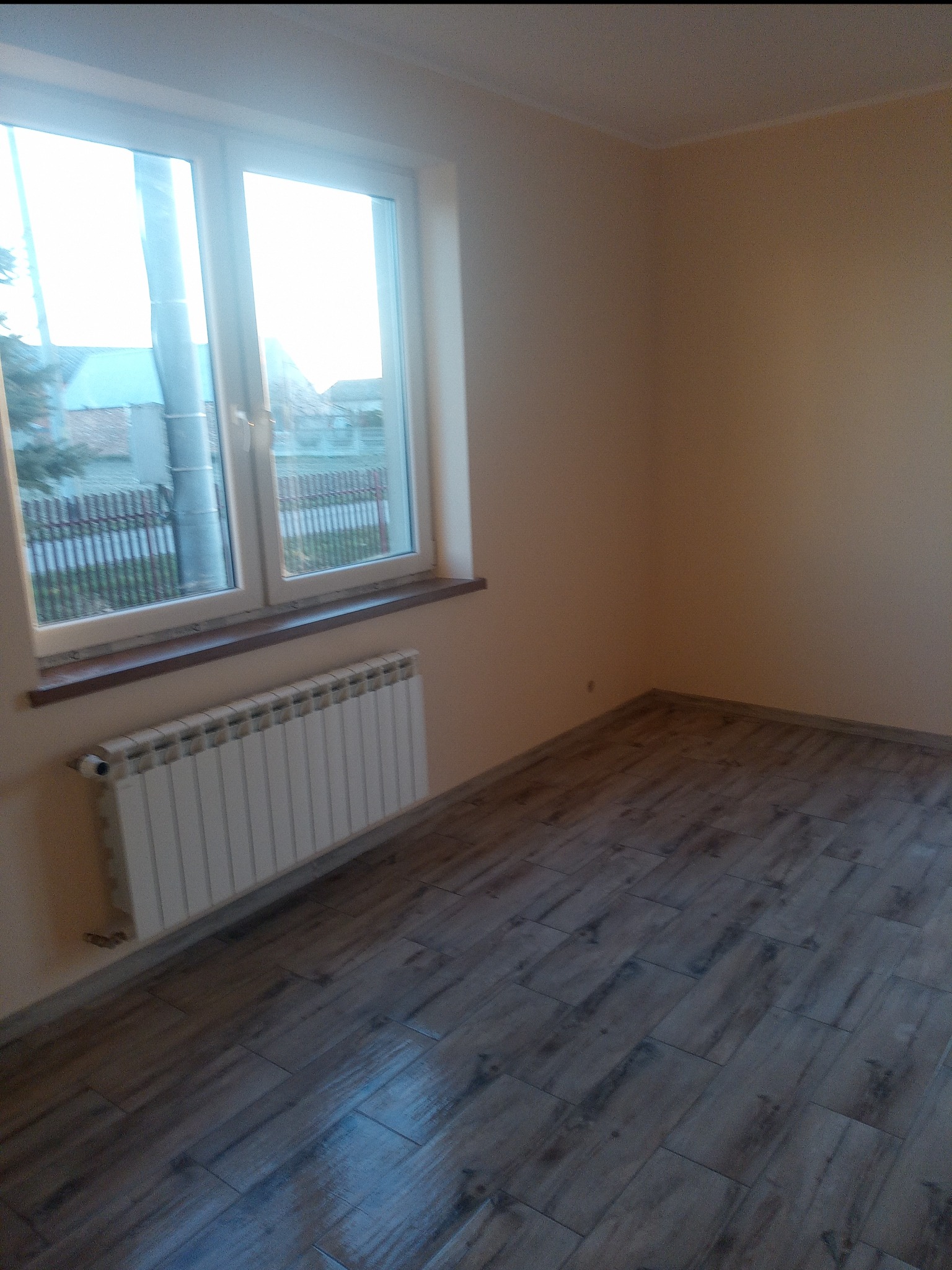 Pusty pokój po remoncie z nową podłogą imitującą drewno, grzejnikiem pod oknem i ścianami w odcieniach beżu.