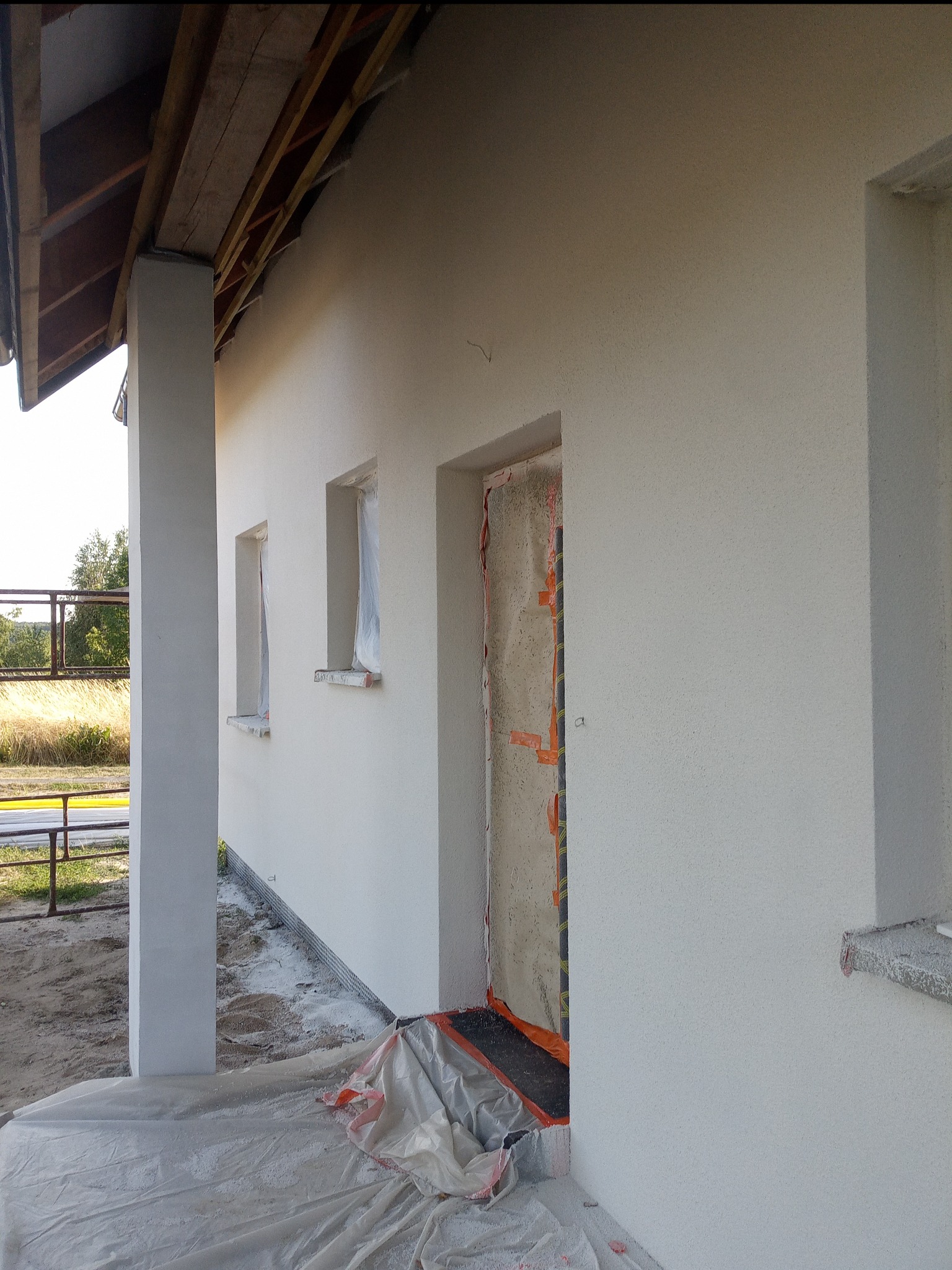 Fragment elewacji domu jednorodzinnego z oknami zabezpieczonymi folią, widoczny fragment konstrukcji dachu i kolumna werandy, teren wokół zabezpieczony folią malarską.