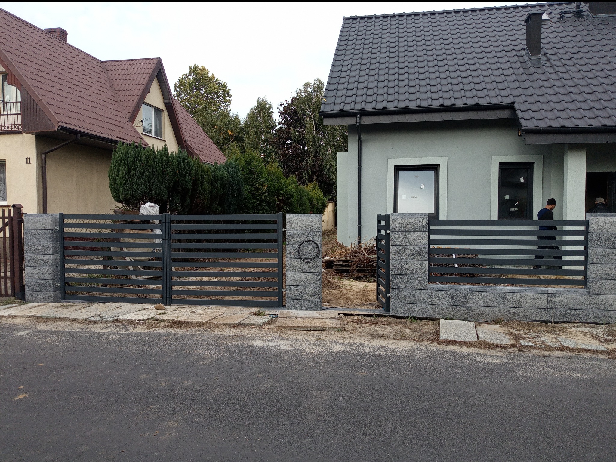 Nowoczesna, pozioma, grafitowa brama wjazdowa z kamiennymi słupkami na tle dwóch domów jednorodzinnych, widok z poziomu ulicy.