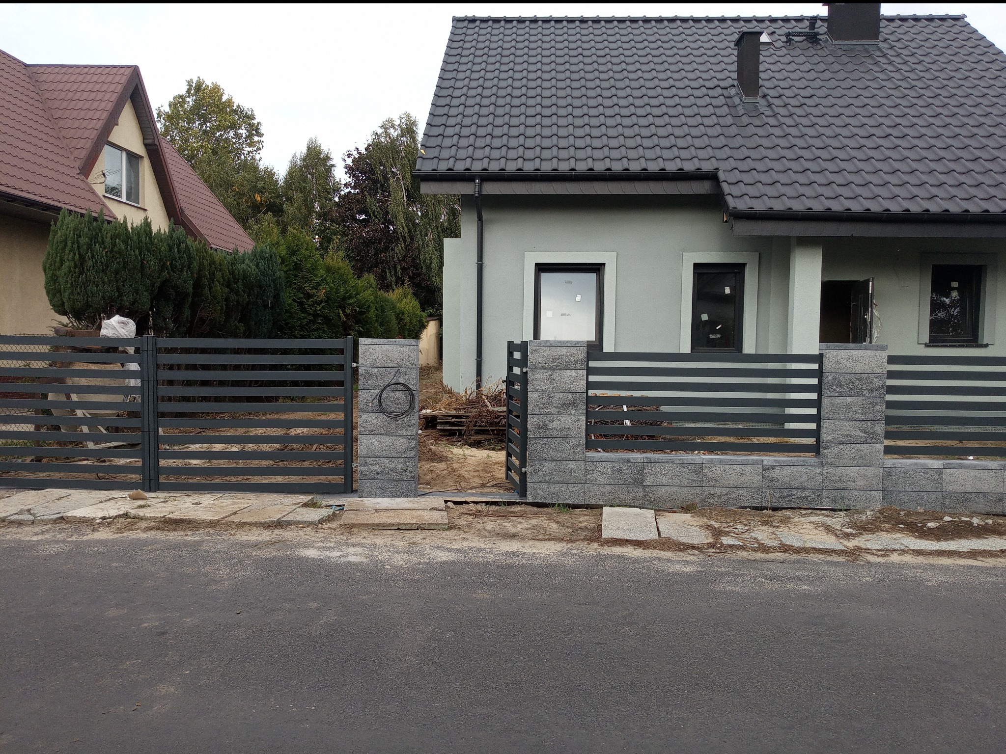 Nowoczesny dom z szarą elewacją i grafitową dachówką, ogrodzony metalowym płotem z elementami z szarego kamienia, widok z ulicy.