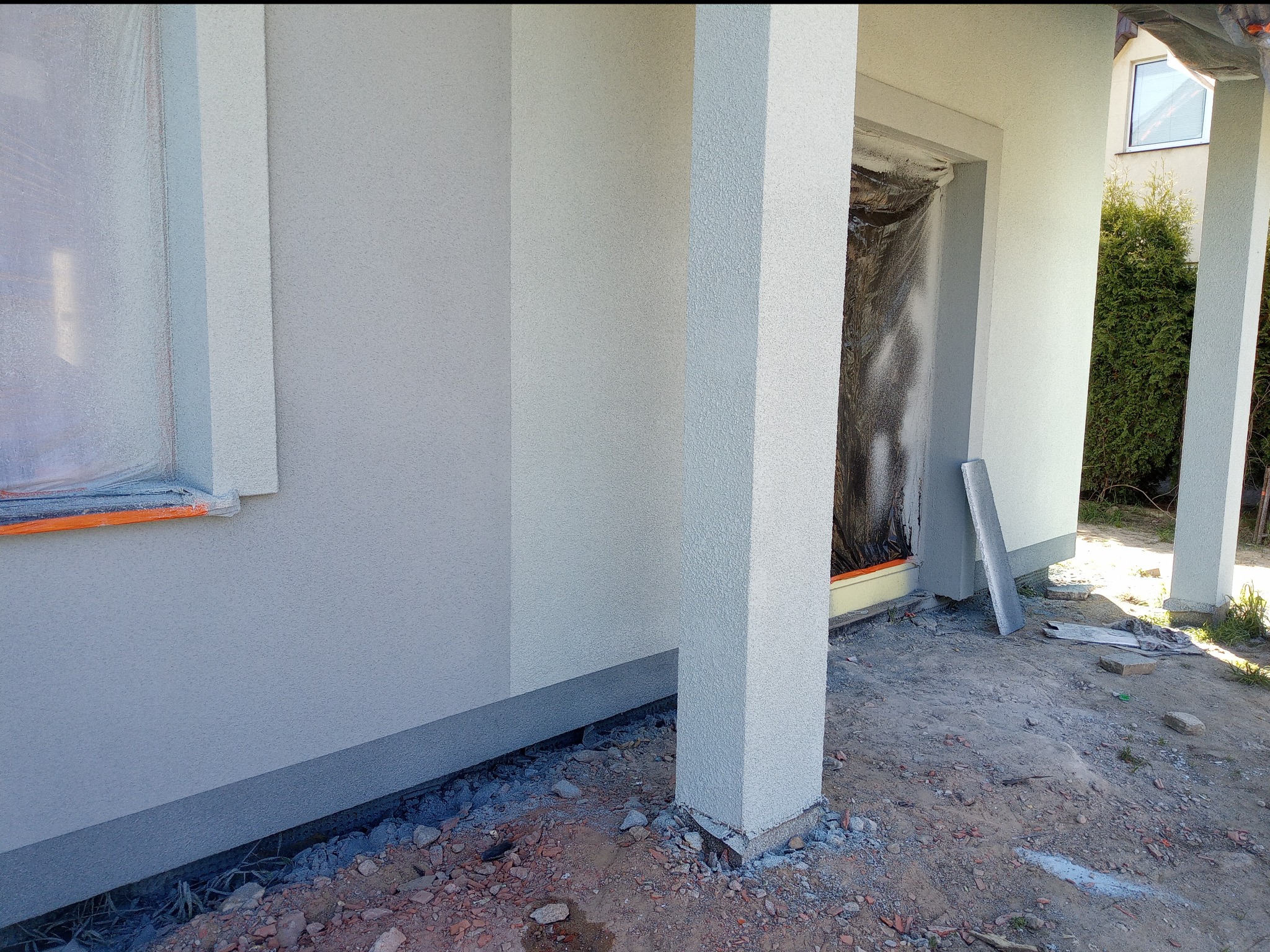 Fragment elewacji budynku w trakcie wykańczania, z oknem zabezpieczonym folią i widocznymi elementami konstrukcyjnymi, teren wokół budynku częściowo utwardzony.