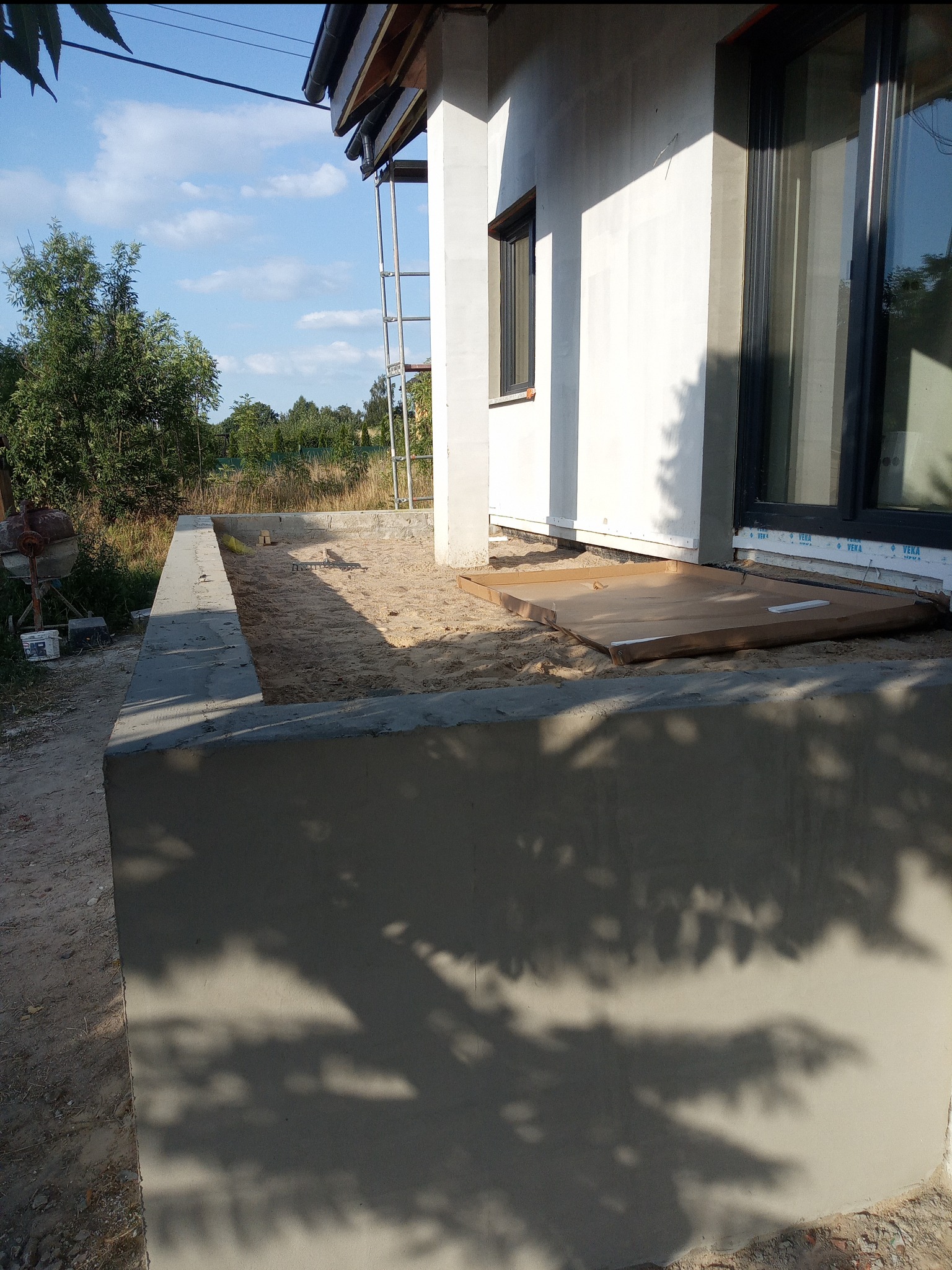 Wczesny etap budowy domu: widoczny fundament z betonu, piasek, ściana budynku z oknem oraz rusztowanie w tle.
