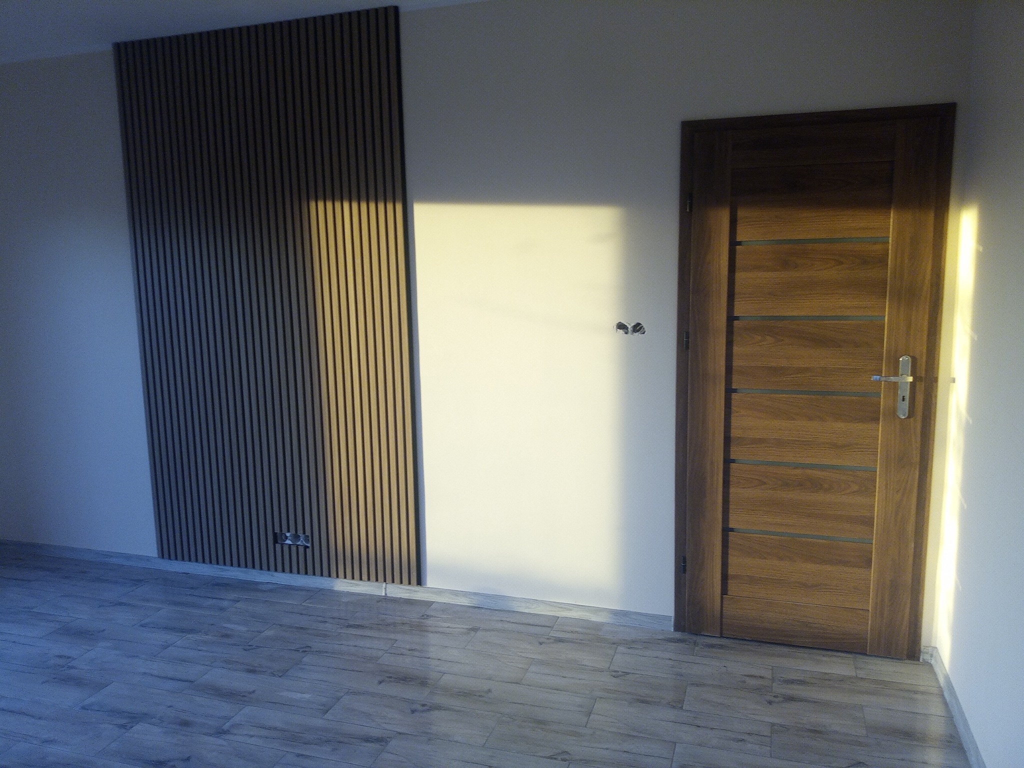 Nowoczesne wnętrze z drewnianą ścianą panelową, drewnianymi drzwiami z poziomymi przeszkleniami i podłogą imitującą drewno, oświetlone popołudniowym słońcem