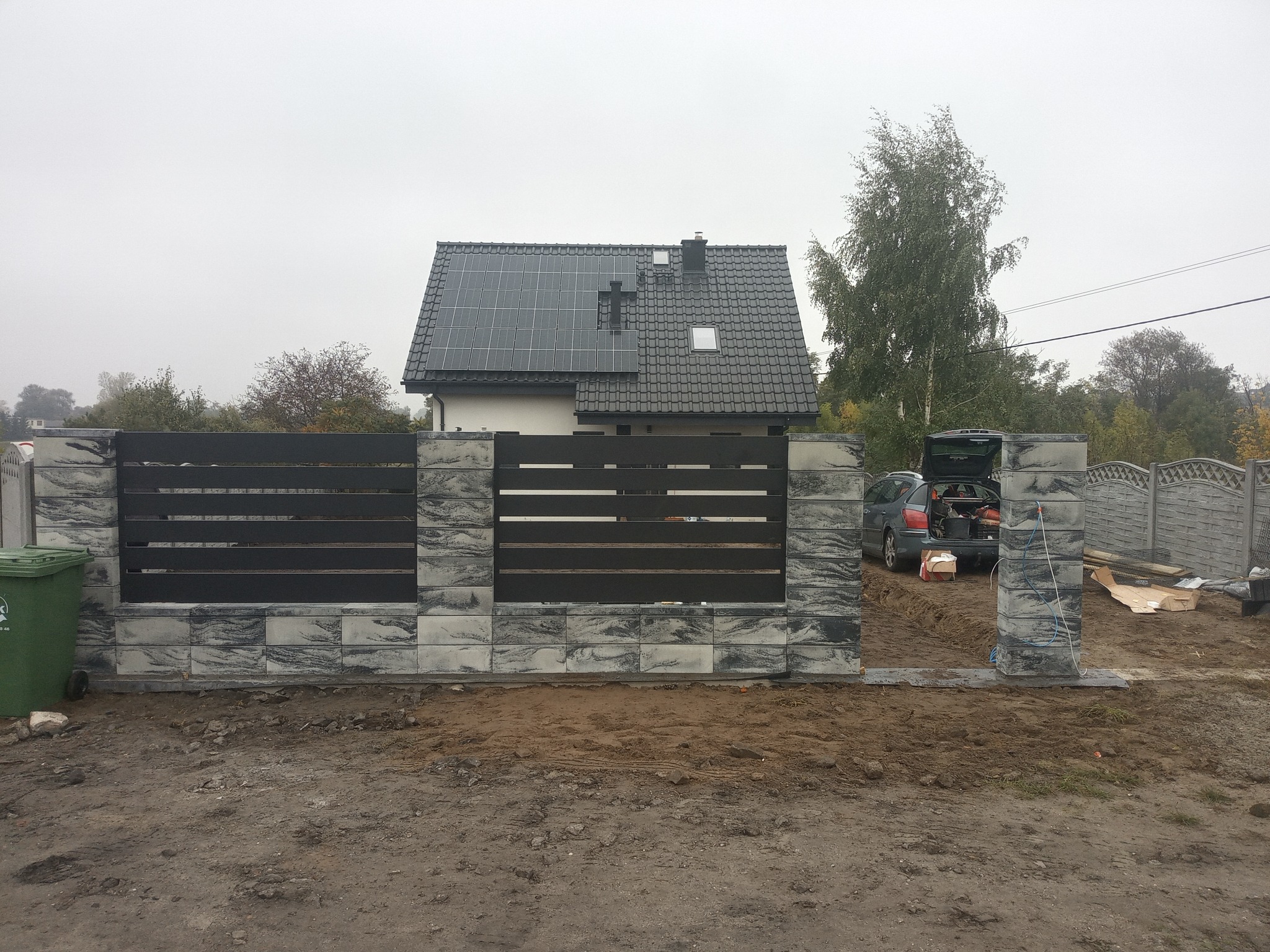 Nowoczesne ogrodzenie z poziomymi, grafitowymi przęsłami i kamiennymi słupkami w odcieniach szarości, na tle domu z panelami słonecznymi na dachu, w trakcie montażu, z widocznym samochodem...