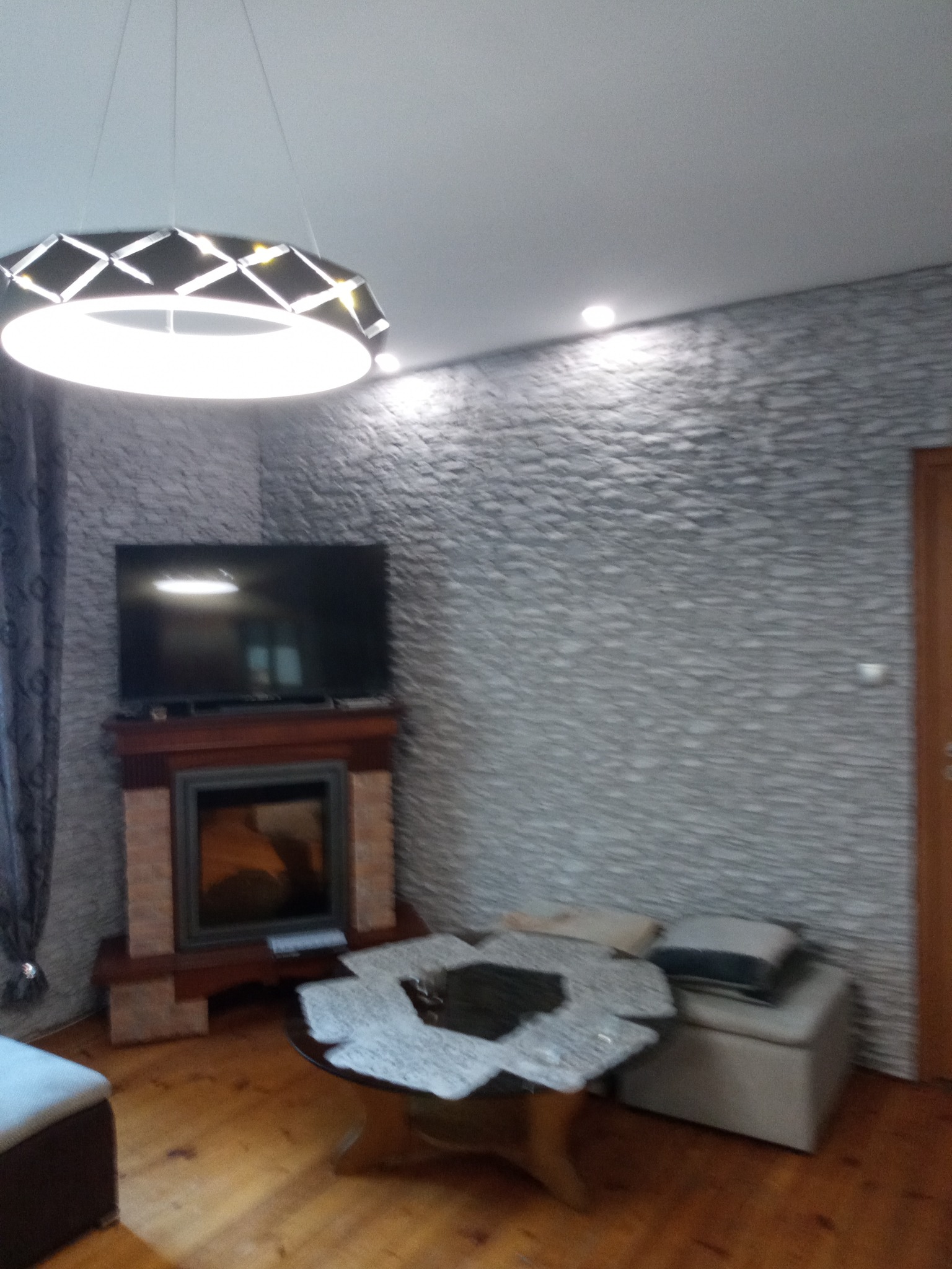 Pokój dzienny z dekoracyjną ścianą z kamienia, kominkiem pod telewizorem i nowoczesnym oświetleniem sufitowym.