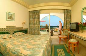 Pokój hotelowy z balkonem i widokiem na morze, Tunezja. W pokoju dwa łóżka z wzorzystą pościelą, telewizor i stolik z krzesłami na balkonie. Na balkonie widoczne osoby i parasol.