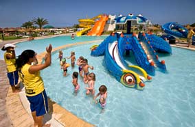 Grupa dzieci plus opiekunowie w płytkim basenie z atrakcjami wodnymi, w tle kolorowe zjeżdżalnie, słoneczny dzień w ośrodku wypoczynkowym.