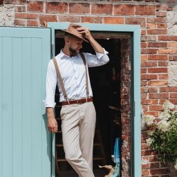 Anastasiia Martynets - Mężczyzna w kapeluszu i szelkach, oparty o turkusowe drzwi w ceglanej ścianie, uśmiecha się, patrząc w dół.
