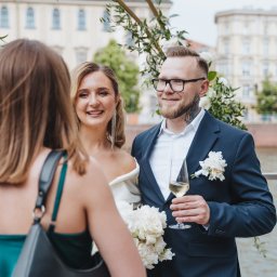 Anastasiia Martynets - Uśmiechnięta panna młoda w białej sukni i pan młody w granatowym garniturze z kieliszkiem szampana, stojący pod dekoracją kwiatową, z widokiem na rzekę i budynki w tle, rozmawiają z kobietą...