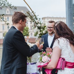 Anastasiia Martynets - Toast weselny na tle miejskiej architektury: para młoda i goście wznoszą kieliszki szampana przy dekorowanej kwiatami konstrukcji.