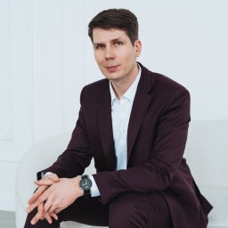 Anastasiia Martynets - Mężczyzna w bordowym garniturze siedzący na jasnej sofie, z zegarkiem na nadgarstku i rękami splecionymi na kolanach na tle białej ściany.