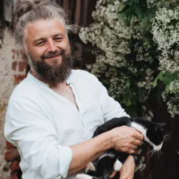 Uśmiechnięty mężczyzna z brodą i włosami spiętymi w kok, trzyma czarno-białego kota na tle drewnianej ściany i kwitnącego krzewu.