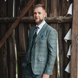 Anastasiia Martynets - Mężczyzna w trzyczęściowym garniturze w kratę pozuje na tle drewnianej konstrukcji z dekoracjami z białych chorągiewek i gipsówki.