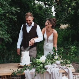 Anastasiia Martynets - Uśmiechnięta para młoda we Wrocławiu kroi tort weselny na drewnianym stole udekorowanym kwiatami i lnem, w tle zielone drzewa.