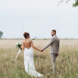 Anastasiia Martynets - Para młoda trzymająca się za ręce idzie przez łąkę w sukni ślubnej z odkrytymi plecami i szarym garniturze w kratę, turbiny wiatrowe w oddali.