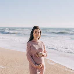 Anastasiia Martynets - Uśmiechnięta kobieta w ciąży, ubrana w różowy komplet, stoi na piaszczystej plaży z falami w tle, trzymając ręce na brzuchu.