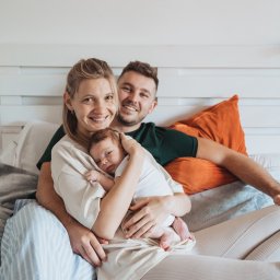 Anastasiia Martynets - Rodzinne ujęcie: uśmiechnięci rodzice tulą noworodka na łóżku z dekoracyjnym łapaczem snów w tle.