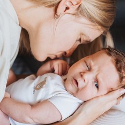 Anastasiia Martynets - Matka pochyla się nad noworodkiem w białym body z nadrukiem misia, delikatne światło, bliski kadr.