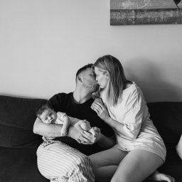Anastasiia Martynets - Czarno-białe zdjęcie przedstawia rodziców siedzących na sofie, całujących się, podczas gdy ojciec trzyma śpiącego noworodka w paski. Na ścianie wisi abstrakcyjny obraz.