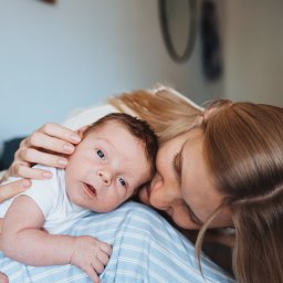 Anastasiia Martynets - Mama przytula noworodka z otwartymi, niebieskimi oczami, leżącego na niebieskiej, pasiastej tkaninie.