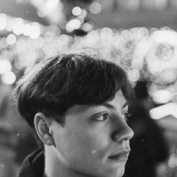 Anastasiia Martynets - Czarno-biały portret młodego mężczyzny z krótkimi włosami, oświetlonego rozmytymi światłami w tle, z widocznymi drobnymi cząsteczkami na włosach.