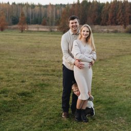 Anastasiia Martynets - Rodzinne zdjęcie na łące: uśmiechnięty mężczyzna obejmuje ciężarną kobietę w kremowej sukience, zza nóg wystaje mały chłopiec.