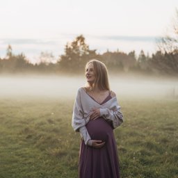 Anastasiia Martynets - Kobieta w ciąży w długiej sukience i swetrze, pozuje na łące we mgle, z zamyślonym wyrazem twarzy, w tle drzewa.