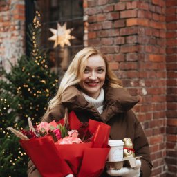 Anastasiia Martynets - Uśmiechnięta blondynka w brązowej kurtce pozuje z bukietem kwiatów w czerwonym papierze i kawą na tle ceglanej ściany i choinki z lampkami.