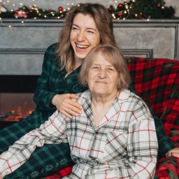 Anastasiia Martynets - Uśmiechnięta młoda kobieta w zielonej piżamie w kratę obejmuje starszą kobietę w białej piżamie w kratę na tle kominka z dekoracjami świątecznymi.