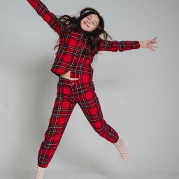 Anastasiia Martynets - Dziewczynka z zamkniętymi oczami w czerwonej piżamie w kratę skacze na jasnoszarym tle, unosząc ręce w górę.