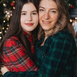 Anastasiia Martynets - Uśmiechnięta matka i córka w koszulach w kratę obejmują się na tle rozświetlonej choinki.