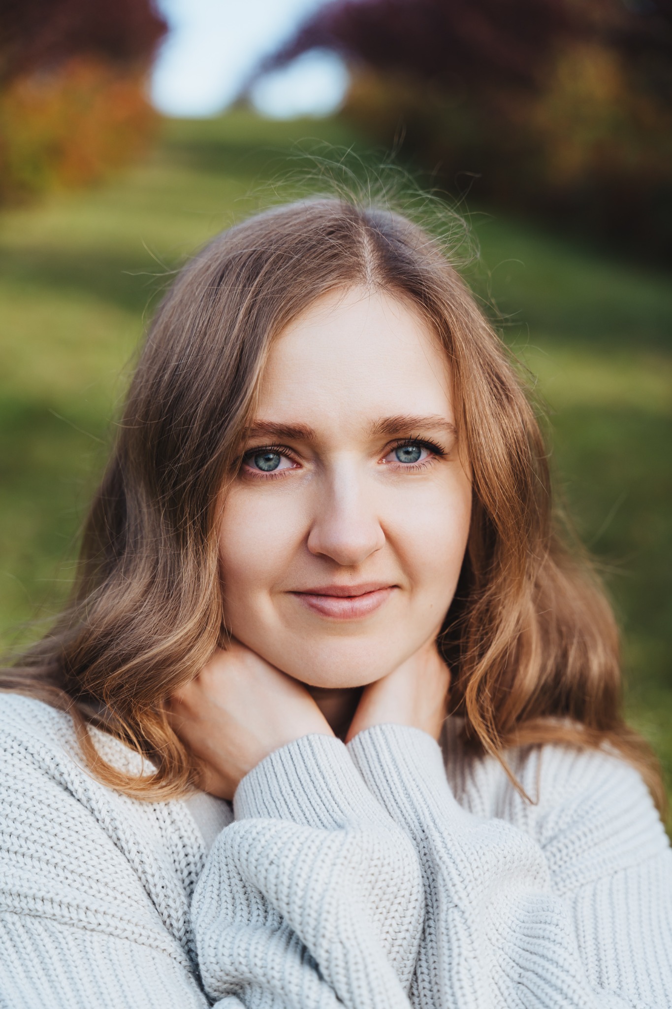 Portret kobiety z długimi blond włosami i niebieskimi oczami, uśmiechniętej, w jasnym swetrze, opierającej podbródek na dłoniach na tle zieleni.