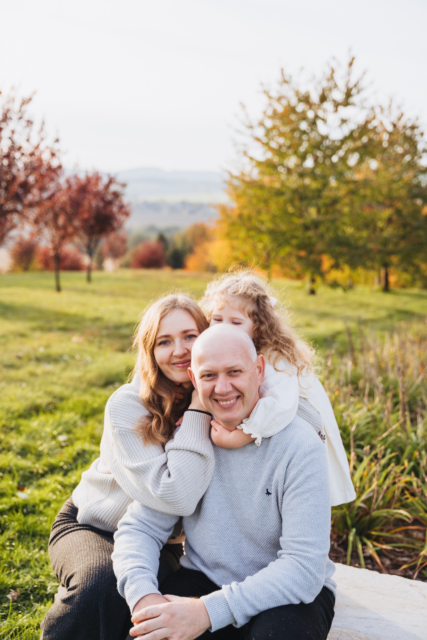 Portret uśmiechniętej rodziny: matka z blond włosami, ojciec i córka z kręconymi włosami, tulący się na tle jesiennego parku.