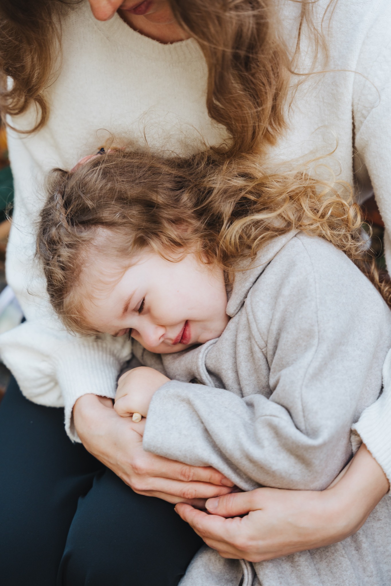 Matka przytula śpiącą córeczkę w szarym płaszczu, widoczne kręcone włosy i delikatny uśmiech dziecka.